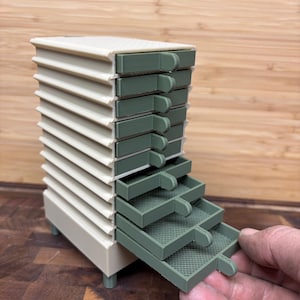 May include: A vertical storage unit with a cream-colored frame and ten stacked drawers in a muted sage green. Each drawer features a small protruding handle on the right side and a textured grid pattern on the interior base. The unit stands on four small legs.