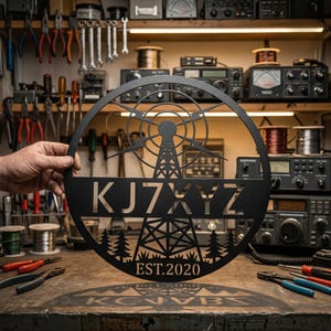Puede incluir: Letrero de metal negro con un diseño circular que presenta una torre de radio, árboles y el texto "KJ7XYZ EST.2020". El letrero se sostiene frente a un banco de trabajo con herramientas y equipos de radio.