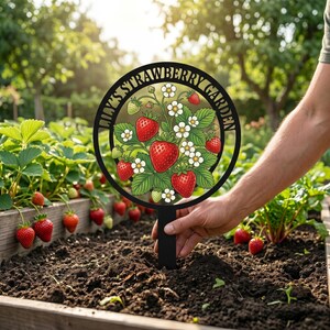 May include: Black metal garden stake with a circular design featuring the text "LILY'S STRAWBERRY GARDEN" around a strawberry illustration. The illustration includes red strawberries, white flowers, and green leaves.