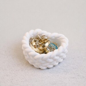 Peut inclure: Un vide-poches blanc en forme de cœur avec un motif de corde tressée. Le plat est rempli de divers bijoux en or et en argent, notamment des bagues, des boucles d'oreilles et des bracelets. Le vide-poches est une solution de rangement décorative.