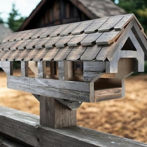 Puede incluir: Un comedero para pájaros de madera diseñado para parecer un puente cubierto, con techo de tejas, paneles laterales abiertos y un poste de soporte central. La estructura está hecha de madera gris desgastada con un acabado rústico y texturizado.