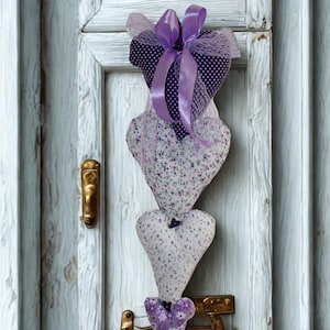 May include: A decorative hanging heart ornament. The ornament features four heart-shaped fabric pieces in shades of purple and white, adorned with floral patterns and polka dots. A purple ribbon and bow complete the design, hanging on a distressed white door.