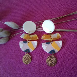 Peut inclure: Une paire de boucles d'oreilles géométriques abstraites. Chaque boucle d'oreille présente un clou circulaire blanc, relié à des formes roses, jaunes et bleu marine. Un charme en spirale doré pend en bas. Les boucles d'oreilles sont posées sur un fond magenta.