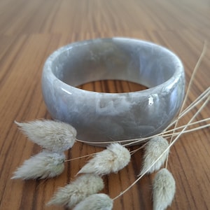 Peut inclure: Un large bracelet jonc circulaire présentant une surface lisse et polie avec un motif marbré dans des tons de gris translucide et de blanc opaque. Le bracelet possède un profil épais et arrondi ainsi qu'une finition brillante et réfléchissante.