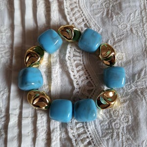 Peut inclure: Un bracelet élastique composé d'une alternance de perles carrées bleu clair à l'aspect marbré et de perles métalliques dorées aux formes irrégulières et réfléchissantes, disposées en cercle pour former une boucle continue.