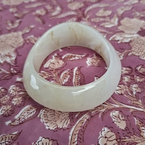 Peut inclure: Un bracelet jonc lisse et translucide de couleur blanc cassé, présentant de subtils motifs marbrés tourbillonnants dans des tons beige et brun clair sur toute sa surface arrondie et polie, offrant un aspect organique et doux.