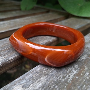 Peut inclure: Un bracelet jonc lisse et poli de forme carrée arrondie. Le bracelet présente une couleur orange brûlé translucide profonde avec des inclusions blanches tourbillonnantes dans tout le matériau, créant un effet marbré sur la surface.