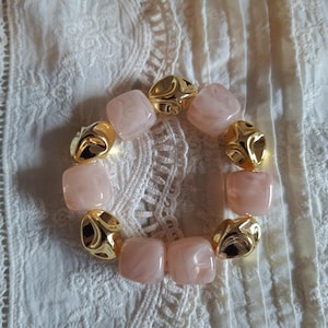 Peut inclure: Un bracelet circulaire composé d'une alternance de perles carrées à la finition marbrée rose pâle et de perles dorées métalliques à la surface facettée et irrégulière. Les perles sont enfilées pour former une boucle aux textures contrastées.