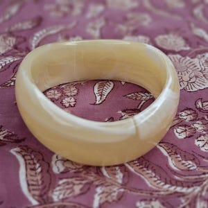 Peut inclure: Un bracelet jonc épais et lisse à l'aspect blanc cassé translucide et crémeux. Cet accessoire circulaire présente de subtils motifs tourbillonnants dans des tons beige clair et foncé sur toute sa surface polie, arrondie et solide.