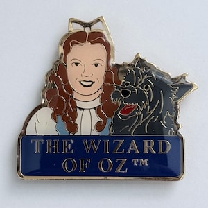 May include: A gold-toned pin featuring a smiling woman with brown hair and a black dog. The woman wears a white blouse and blue dress. The pin has a blue banner with the text "THE WIZARD OF OZ" in gold letters.