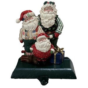 May include: A decorative stocking holder featuring three Santa Claus figures of varying sizes. The figures wear red, white, and green holiday attire, including patterned sweaters and hats. A small blue gift box with a gold ribbon sits at their feet.