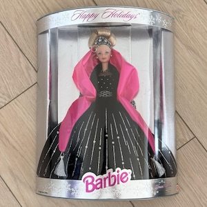 May include: A Barbie doll in a clear display case. The doll wears a black gown with silver accents and a pink shawl. The case is silver with the words "Happy Holidays" at the top and the Barbie logo at the bottom.