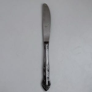 May include: A stainless steel butter knife with a serrated blade and ornate handle. The handle features a detailed floral design. The knife is designed for spreading butter or other soft foods.