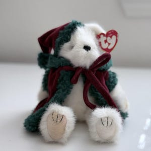 May include: A white plush bear with a red heart-shaped tag. The bear is wearing a dark green and red outfit with a matching bow. The bear's paws are white with tan pads.