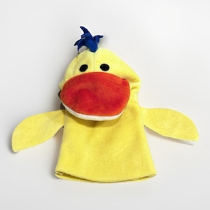 May include: A yellow duck hand puppet with a red beak, black eyes, and a blue crest. The puppet has attached wings and is made of soft fabric. The puppet is designed to fit over the hand for play.