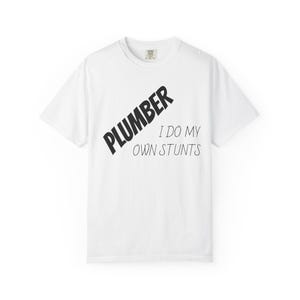 May include: White t-shirt featuring the word "PLUMBER" in bold, black, angled letters. Below, the text reads "I DO MY OWN STUNTS" in a handwritten style. The shirt has a crew neck.