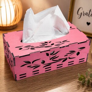 Pode incluir: Uma caixa retangular cor-de-rosa para lenços de papel com padrões de folhas e ramos pretos cortados a laser nas laterais e no topo. A caixa é construída com juntas de encaixe visíveis nos cantos e contém lenços de papel brancos.