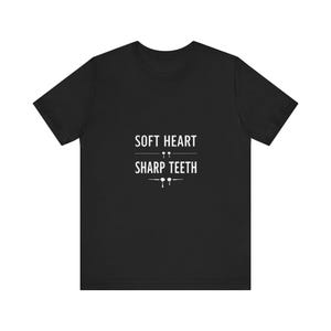May include: Black t-shirt with the white text "SOFT HEART SHARP TEETH". The text is centered and stacked, with small graphic elements above and below the words. The shirt has short sleeves and a crew neck.