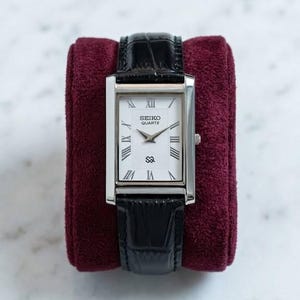 Puede incluir: Un reloj Seiko Quartz rectangular con caja plateada y esfera blanca con números romanos. El reloj tiene una correa de cuero negro y se exhibe sobre una almohada de terciopelo burdeos.