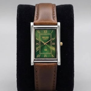 Puede incluir: Un reloj Seiko Quartz rectangular con esfera verde y agujas doradas. El reloj tiene una caja plateada y una correa de cuero marrón. El reloj se muestra sobre una superficie negra.