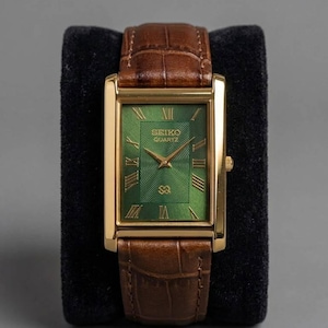 Könnte beinhalten: Eine rechteckige, goldfarbene Seiko Quartz-Uhr mit grünem Zifferblatt und braunem Lederarmband. Das Zifferblatt der Uhr hat römische Ziffern und goldene Zeiger. Die Uhr wird auf einer schwarzen Samtoberfläche präsentiert.