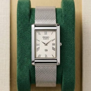 Pode incluir: Um relógio Seiko Quartz retangular com pulseira de malha prateada e mostrador creme. O relógio apresenta algarismos romanos como marcadores de hora. O relógio é exibido em um suporte de veludo verde.