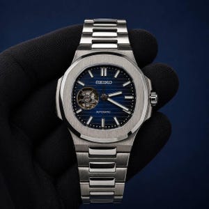 Puede incluir: Un reloj de pulsera automático Seiko de acero inoxidable con esfera azul de rayas horizontales, volante visible a las nueve, índices de bastón plateados y una pulsera de eslabones de metal cepillado integrada a juego.