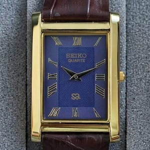 Op de afbeelding: Een rechthoekig, goudkleurig Seiko Quartz horloge met een blauwe wijzerplaat en bruine leren band. De wijzerplaat van het horloge heeft gouden Romeinse cijfers en wijzers. Het horloge wordt gepresenteerd in een grijze doos.
