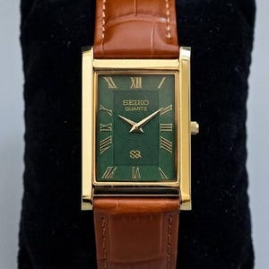 May include: A rectangular gold-toned Seiko Quartz watch with a green face and Roman numeral hour markers. The watch has gold hands and a brown leather band. The watch is displayed on a black velvet surface.
