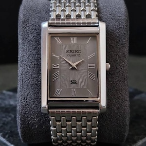 May include: A rectangular Seiko Quartz watch with a silver-tone case and band. The gray dial features Roman numeral hour markers and the Seiko logo. The watch is displayed on a gray velvet surface.
