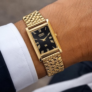 May include: A gold-tone rectangular wristwatch with a black dial, featuring gold Roman numeral hour markers and gold hands. The watch has a textured gold-tone metal link bracelet and displays the text SEIKO QUARTZ on the watch face.