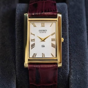 May include: A rectangular gold-toned Seiko Quartz watch with a white face and Roman numeral hour markers. The watch has gold hands and a burgundy leather band. The watch face displays the Seiko Quartz logo and a small symbol. The watch is displayed on a dark blue surface.