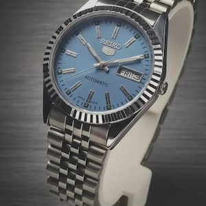 May include: A Seiko 5 automatic wristwatch featuring a light blue dial with silver-tone hour markers and hands. The watch has a stainless steel fluted bezel and a multi-link metal bracelet. A day-date window is positioned at the three o'clock mark.