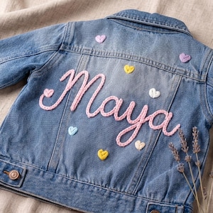 May include: A light blue denim jacket with the name "Maya" embroidered in pink script on the back. The jacket is decorated with small, colorful heart-shaped patches. Gold-colored shoes are visible in the bottom right corner.