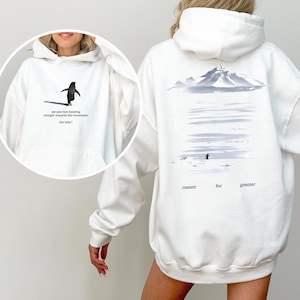May include: White hoodie featuring a penguin graphic on the front with the text "we saw him heading straight towards the mountains but why?". The back of the hoodie has a mountain scene and the words "meant for greater".
