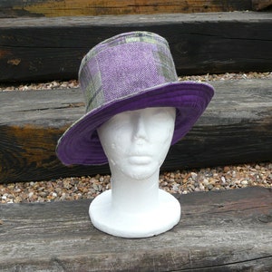 May include: A reversible bucket hat with a purple and green plaid exterior and a solid purple interior. The hat features a herringbone pattern on the crown. It is displayed on a white mannequin head. The hat is made of a textured fabric.