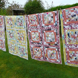 May include: Four colourful patchwork quilts are hanging on a washing line outdoors. The quilts are made from a variety of fabrics in shades of red, pink, yellow and white. The quilts are all different sizes and have a variety of patterns.