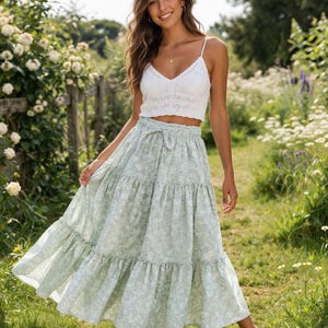May include: A white crochet crop top with spaghetti straps and a V-neck, worn with a light green floral print midi skirt. The skirt features a tiered design and a matching tie belt. The outfit is worn outdoors in a garden setting.