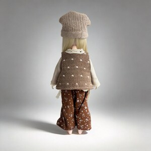 May include: A doll wearing a knitted brown vest with white polka dots, a beige long-sleeved shirt, and brown floral pants. The doll also has a matching brown knitted hat. The doll's blonde hair is visible.