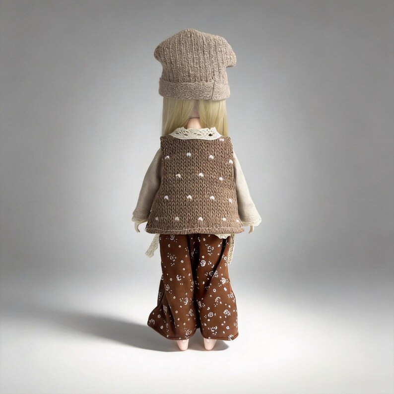 May include: A doll wearing a knitted brown vest with white polka dots, a beige long-sleeved shirt, and brown floral pants. The doll also has a matching brown knitted hat. The doll's blonde hair is visible.