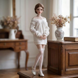 Puede incluir: Una mu&ntilde;eca articulada con cabello casta&ntilde;o recogido, que viste un vestido tipo su&eacute;ter blanco de punto trenzado con hombros descubiertos, medias altas de encaje blanco y zapatos tipo Mary Jane blancos con una tira y una peque&ntilde;a hebilla.