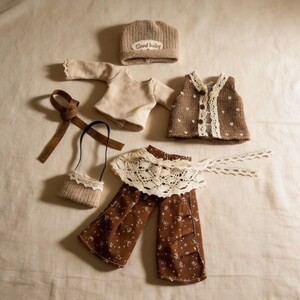 May include: A doll clothing set featuring a beige knit hat with "Good baby" text, a beige long-sleeve shirt, a brown vest with white heart accents, brown floral pants, a small purse, and a brown ribbon.
