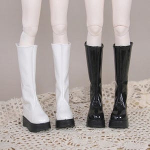 May include: Two pairs of knee-high doll boots with platform soles. One pair is glossy white and the other pair is glossy black. Each boot features a vertical seam running up the front center and a thick, solid black platform base for the doll feet.