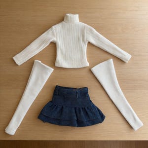 May include: A doll clothing set featuring a cream-colored ribbed turtleneck sweater, a denim skirt with a ruffled hem, and two white leg warmers. The items are arranged on a light brown wooden surface.
