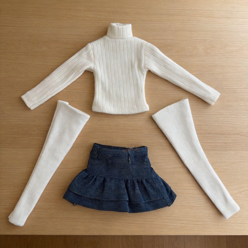 May include: A doll clothing set featuring a cream-colored ribbed turtleneck sweater, a denim skirt with a ruffled hem, and two white leg warmers. The items are arranged on a light brown wooden surface.