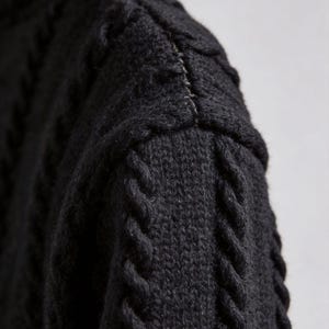 Pu&ograve; includere: Primo piano di un maglione nero a maglia con motivo a trecce. La cucitura della spalla &egrave; visibile, con una cucitura di colore chiaro. La trama della lana &egrave; evidente, mostrando l'intricato motivo e la costruzione del capo.