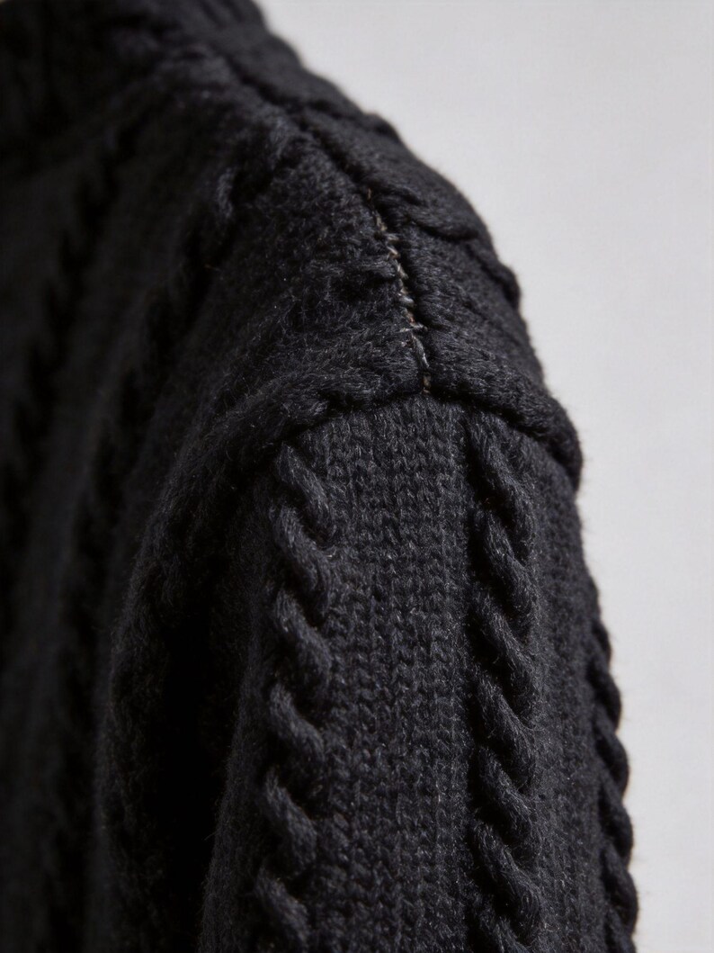 Pu&ograve; includere: Primo piano di un maglione nero a maglia con motivo a trecce. La cucitura della spalla &egrave; visibile, con una cucitura di colore chiaro. La trama della lana &egrave; evidente, mostrando l'intricato motivo e la costruzione del capo.