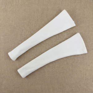 May include: Two white, ribbed knit arm warmers are displayed against a neutral background. The arm warmers are long, with a tapered design, and appear to be made of a soft, textured fabric. They are likely designed to be worn on the arms for warmth or fashion.