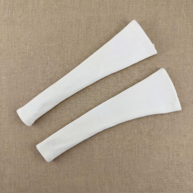 May include: Two white, ribbed knit arm warmers are displayed against a neutral background. The arm warmers are long, with a tapered design, and appear to be made of a soft, textured fabric. They are likely designed to be worn on the arms for warmth or fashion.