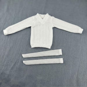 Puede incluir: Un su&eacute;ter blanco de manga larga con cuello en V y un patr&oacute;n de tejido de cable vertical, acompa&ntilde;ado por dos piezas de tela rectangulares blancas separadas con un dise&ntilde;o de malla de tejido abierto texturizado colocado debajo del su&eacute;ter.